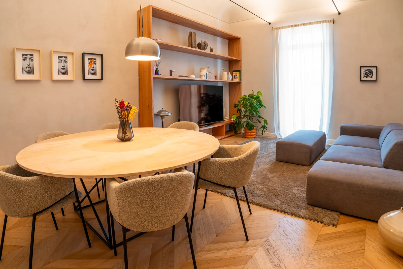 Soggiorno con tavolo tondo in legno, sedie beige, divano grigio e libreria con TV