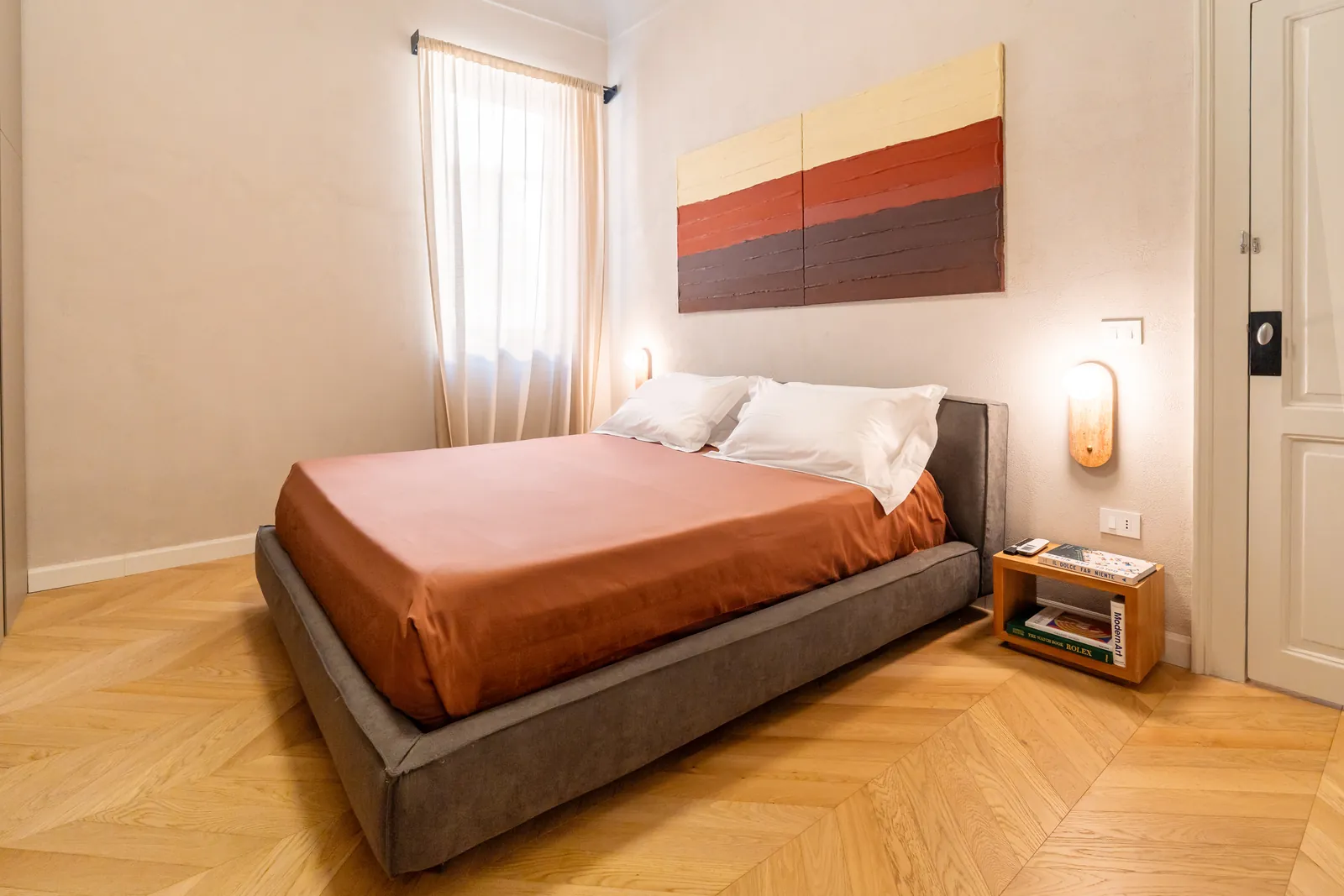 Letto matrimoniale con lenzuola terracotta, lampade in legno e quadro a fasce